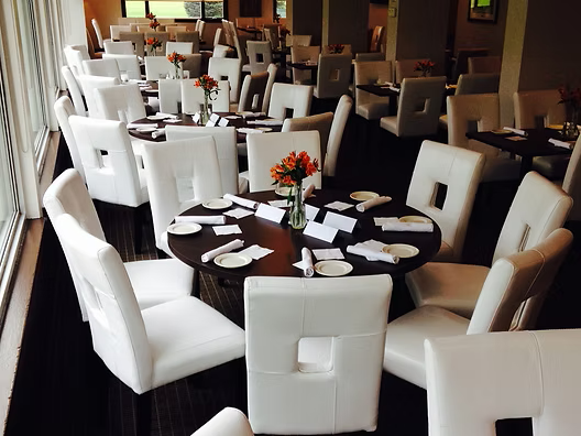 Restaurant dining room with round tables, white chairs, and orange flower centerpieces.