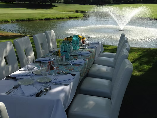 Outdoor table set for a meal, overlooking a pond with a fountain; white chairs, blue accents.