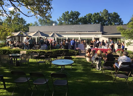 Outdoor gathering with people socializing in front of a white building with a gray roof; tables and chairs on the grass.