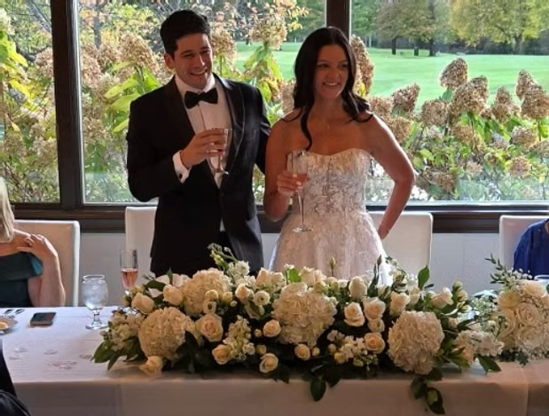 Bride and groom toast with champagne at a wedding reception. White floral centerpiece on table.