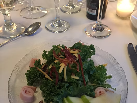 Salad on clear plate with kale, apple slices, ham, and wine glasses.