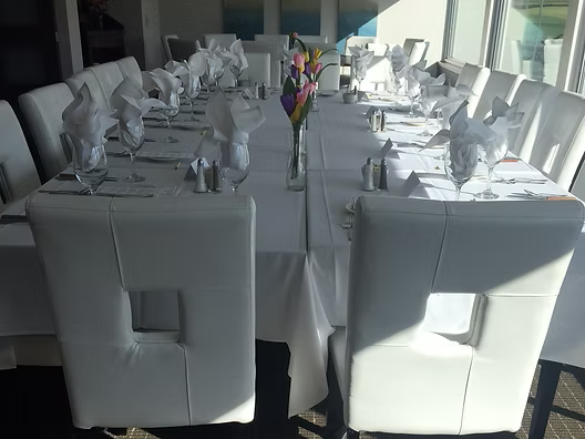 Long white table set for a formal event, with matching chairs, napkins, and a vase of flowers.