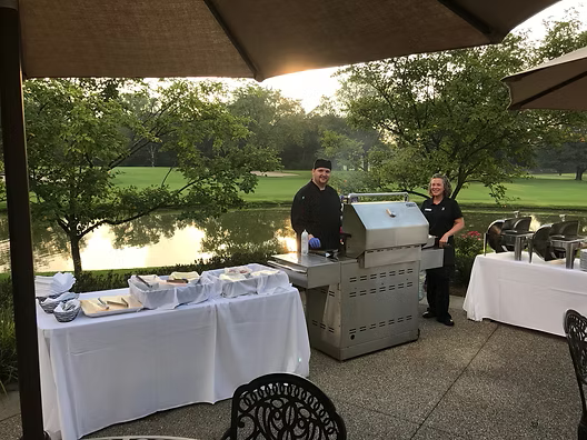 Chefs grilling at an outdoor event near a pond, with food tables set up.