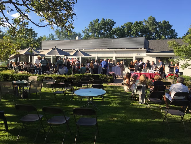 Outdoor gathering with people in front of a white building with a red table, on a sunny day.
