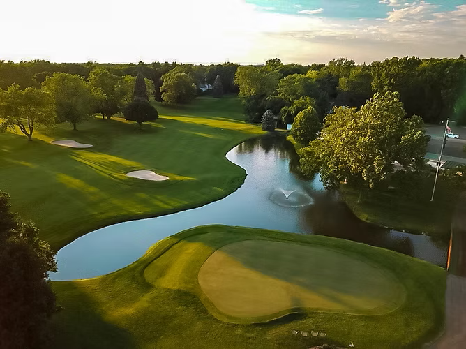Aerial view of a green golf course with a pond, trees, and sand traps.