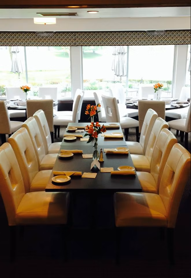 Restaurant dining area with long tables, cream chairs, large windows, and flower centerpieces.