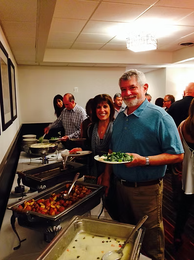 People at a buffet. Man in teal shirt smiles, holding plate of food. Others serve themselves.