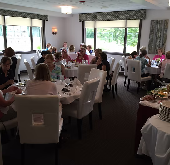 People seated at tables in a brightly lit dining room with windows, possibly at a luncheon or event.