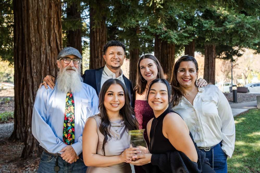 Group of six people posing outdoors by trees. Smiling faces, formal attire, holding an object.