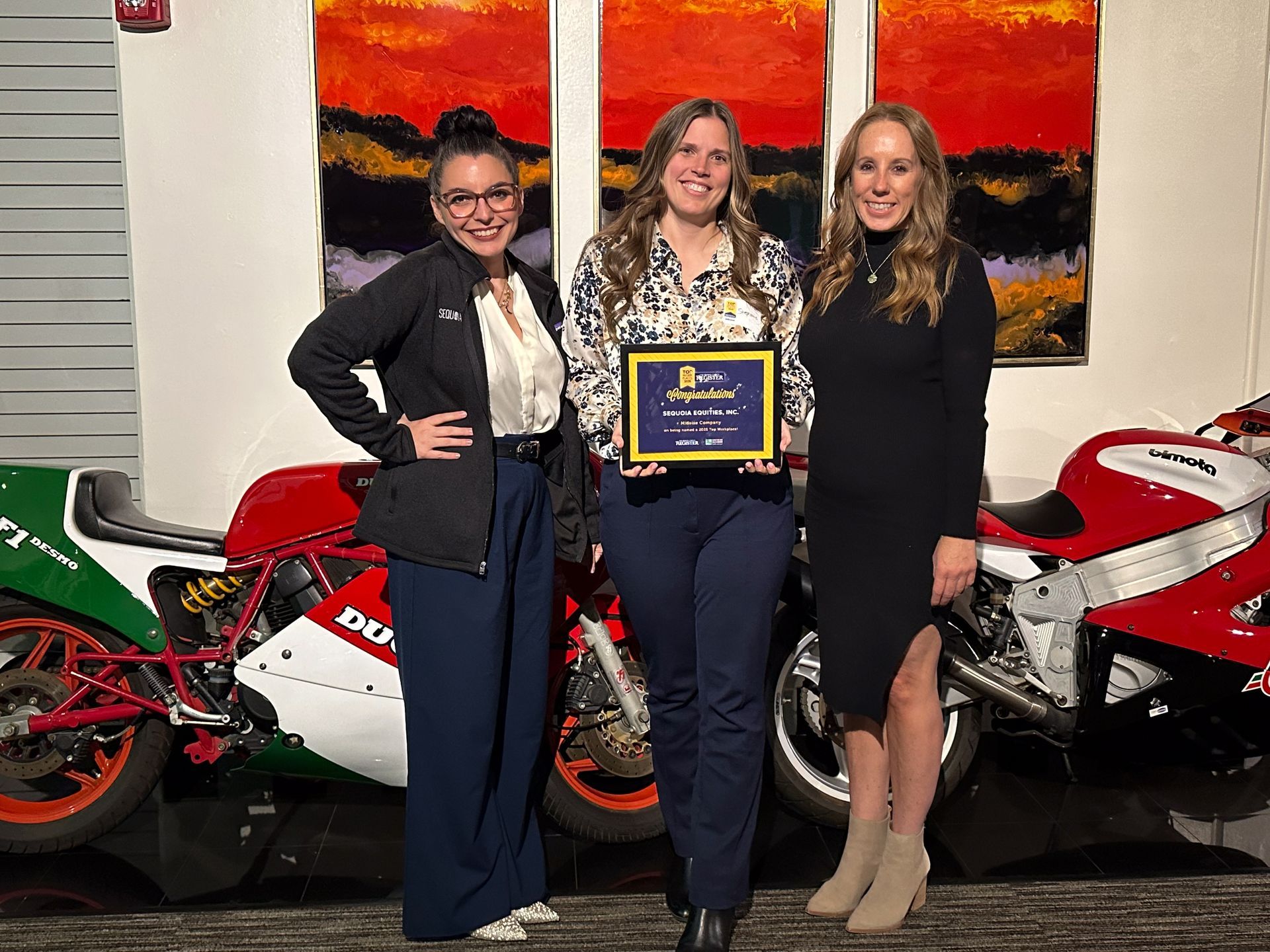 Three women hold an award, standing next to motorcycles and art.