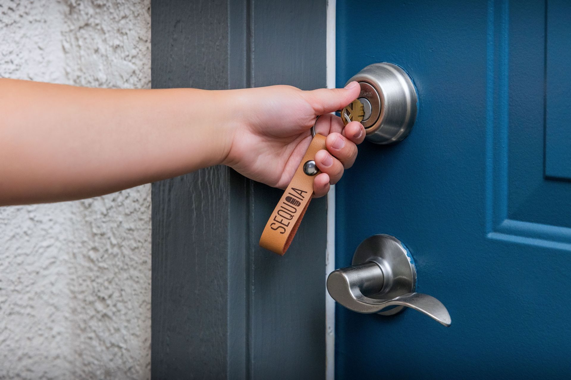 Hand turning a key in a silver door lock on a blue door, with a tan leather keychain.