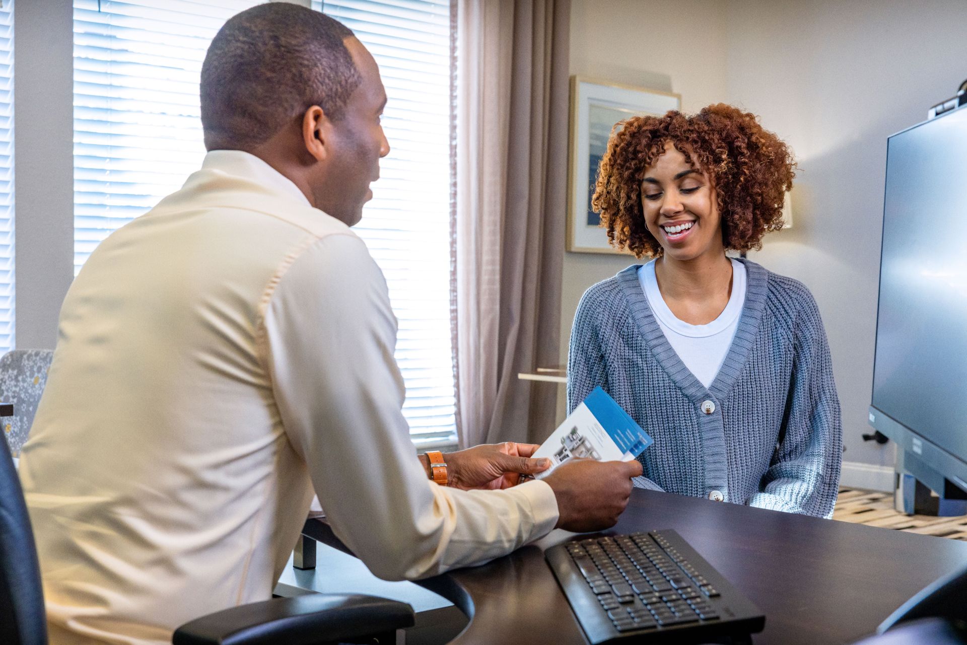 Man in tan shirt showing papers to a smiling woman at a desk in an office setting.