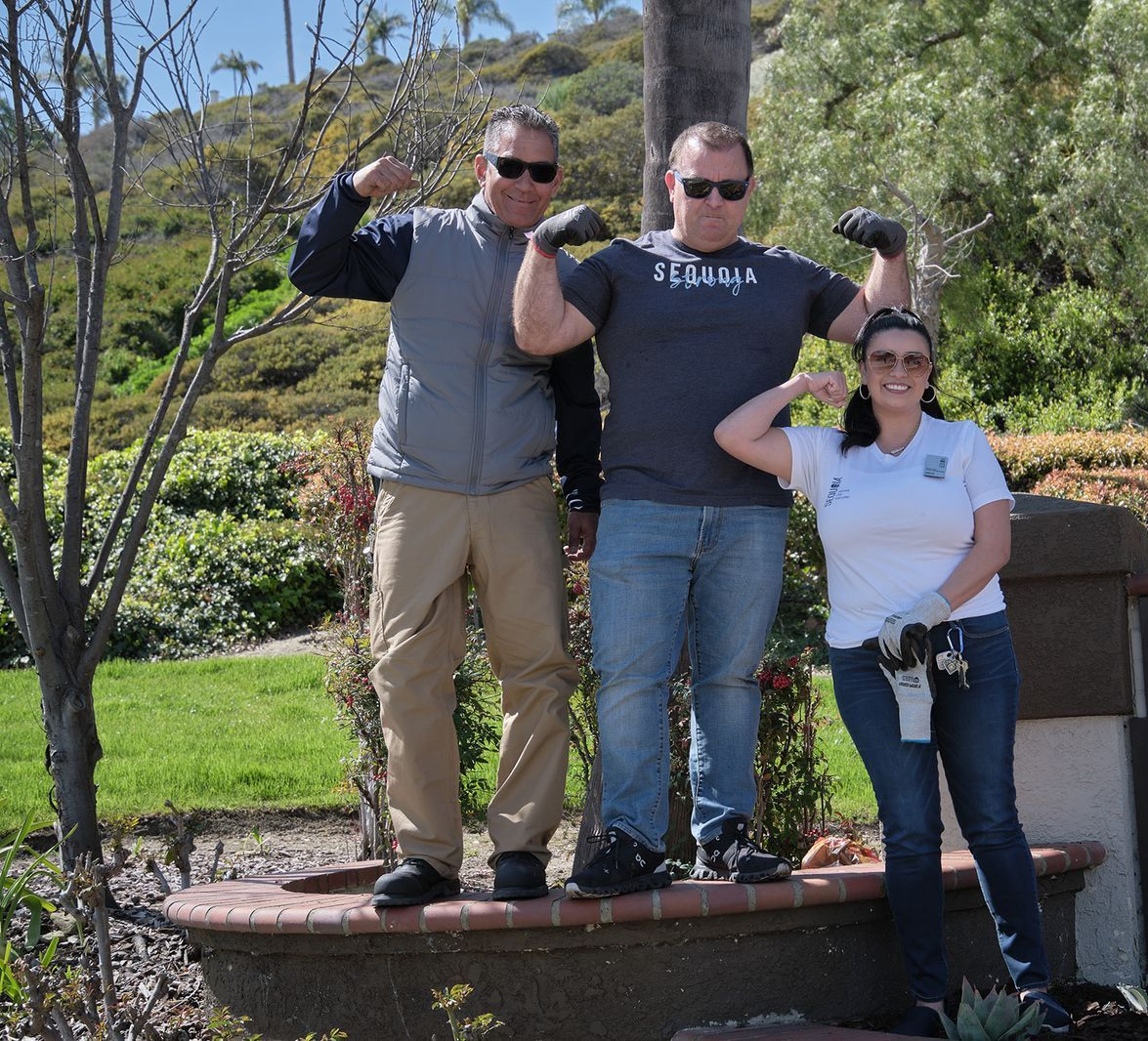 Three people flexing their arms outdoors near landscaping. Two men and one woman smiling.