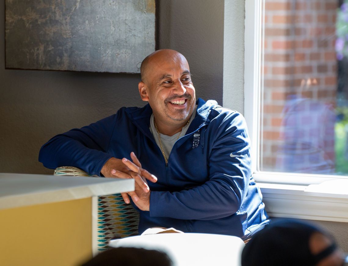 Man in blue jacket smiles, seated near a window, with brick and natural light visible.