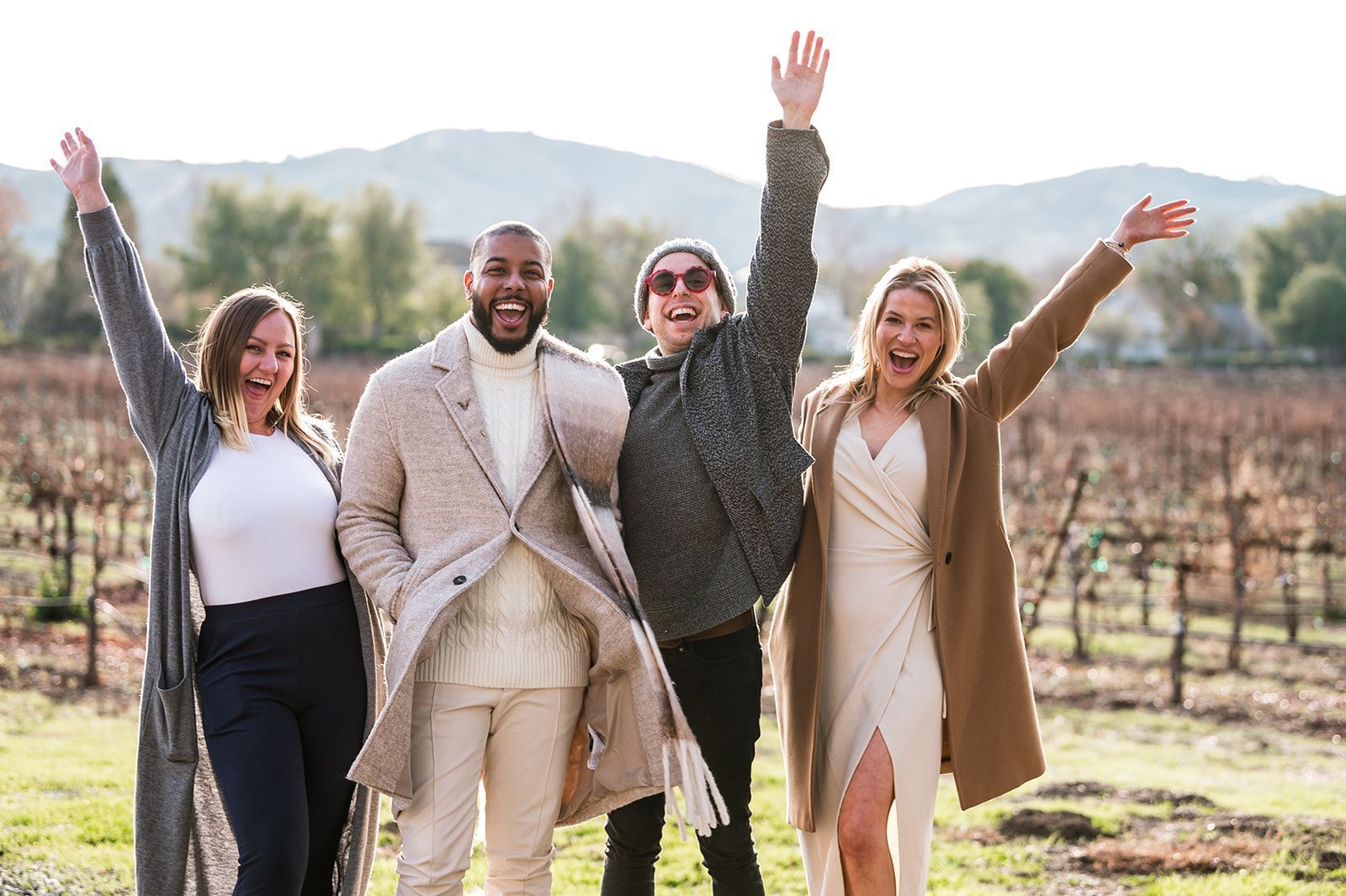 Four people pose outdoors, vineyard background. Two raise arms, all smiling, wearing winter clothing.