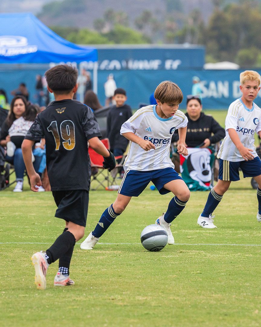 Soccer player in burgundy kicks the ball, another player defends; green field, outdoor game.