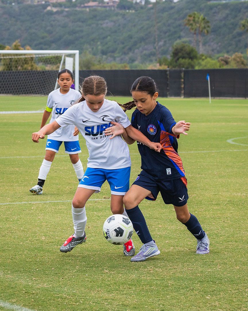 Youth soccer players in action on a green field. One in white kicks the ball as two in blue watch.