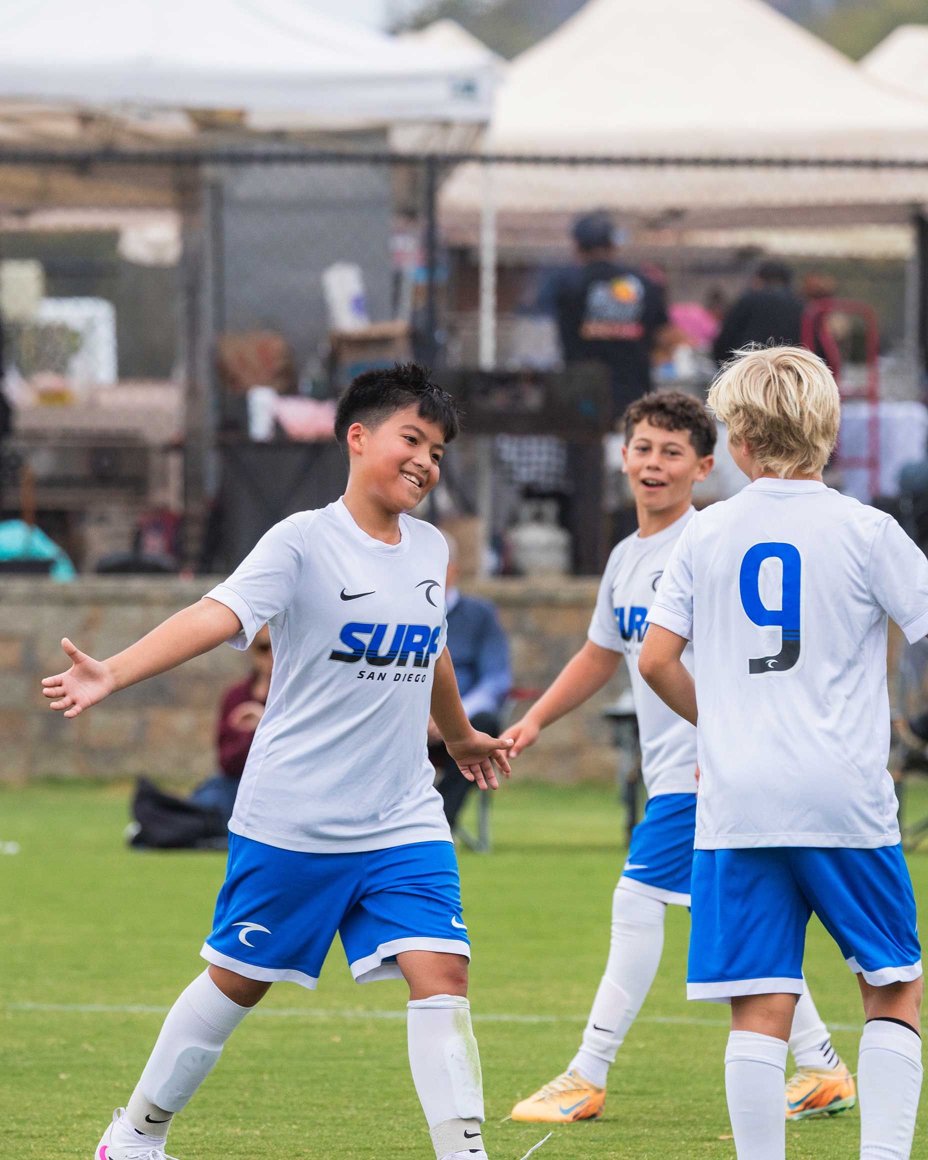 Youth soccer players in white and blue uniforms celebrate on a grassy field during a match