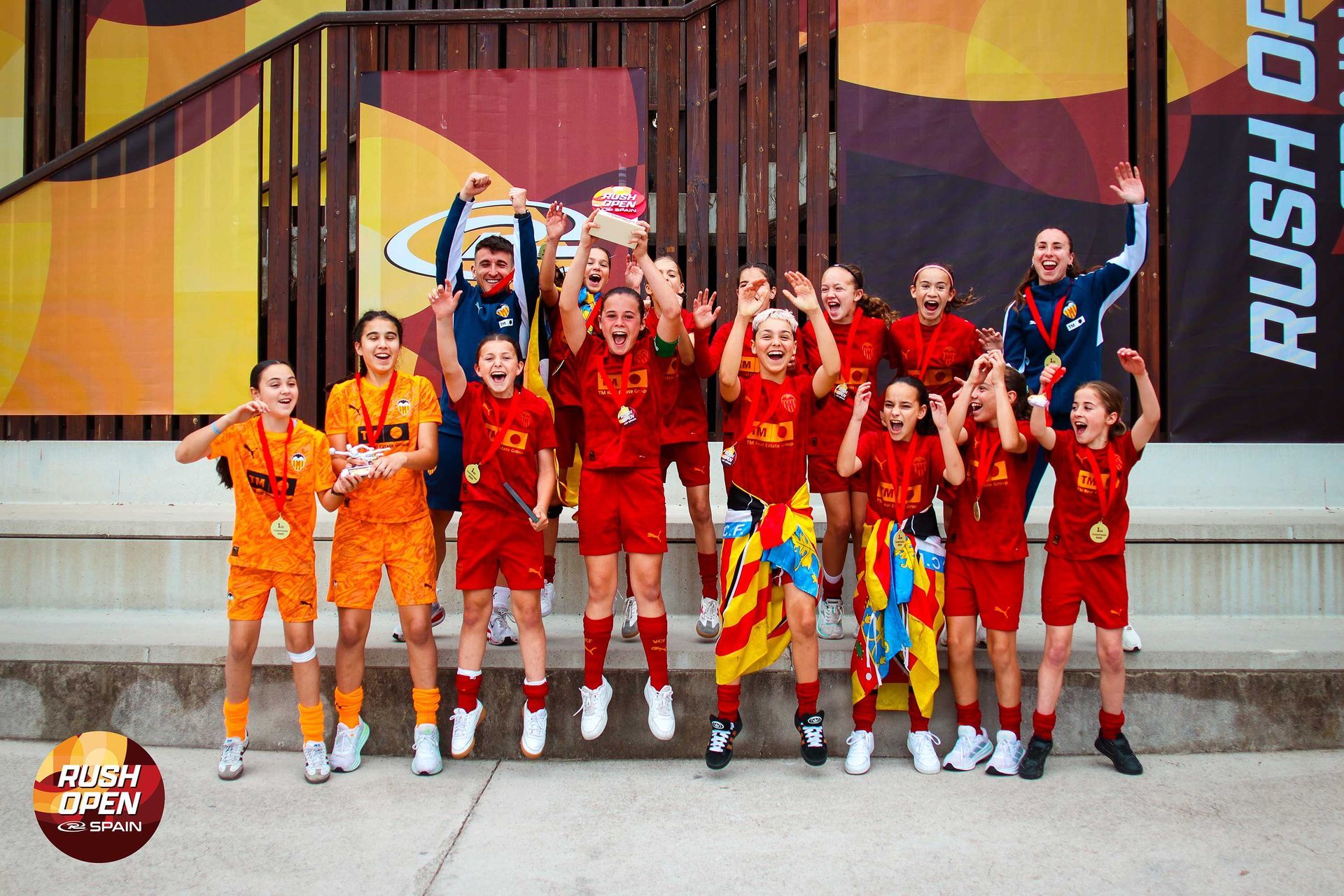 Team in red and orange uniforms posing with raised arms on a stage, with a “RUSH” banner behind them.