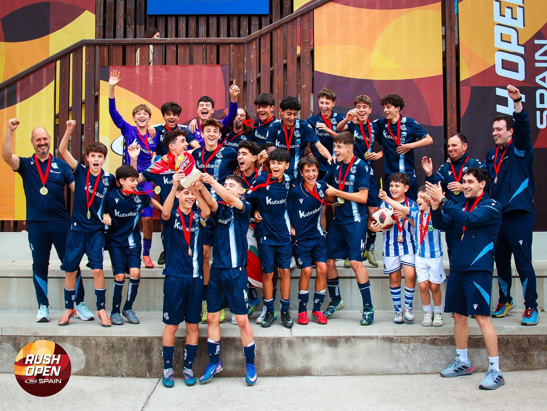 Youth sports team posing on stadium steps, wearing navy jerseys and medals, with a coach at right.