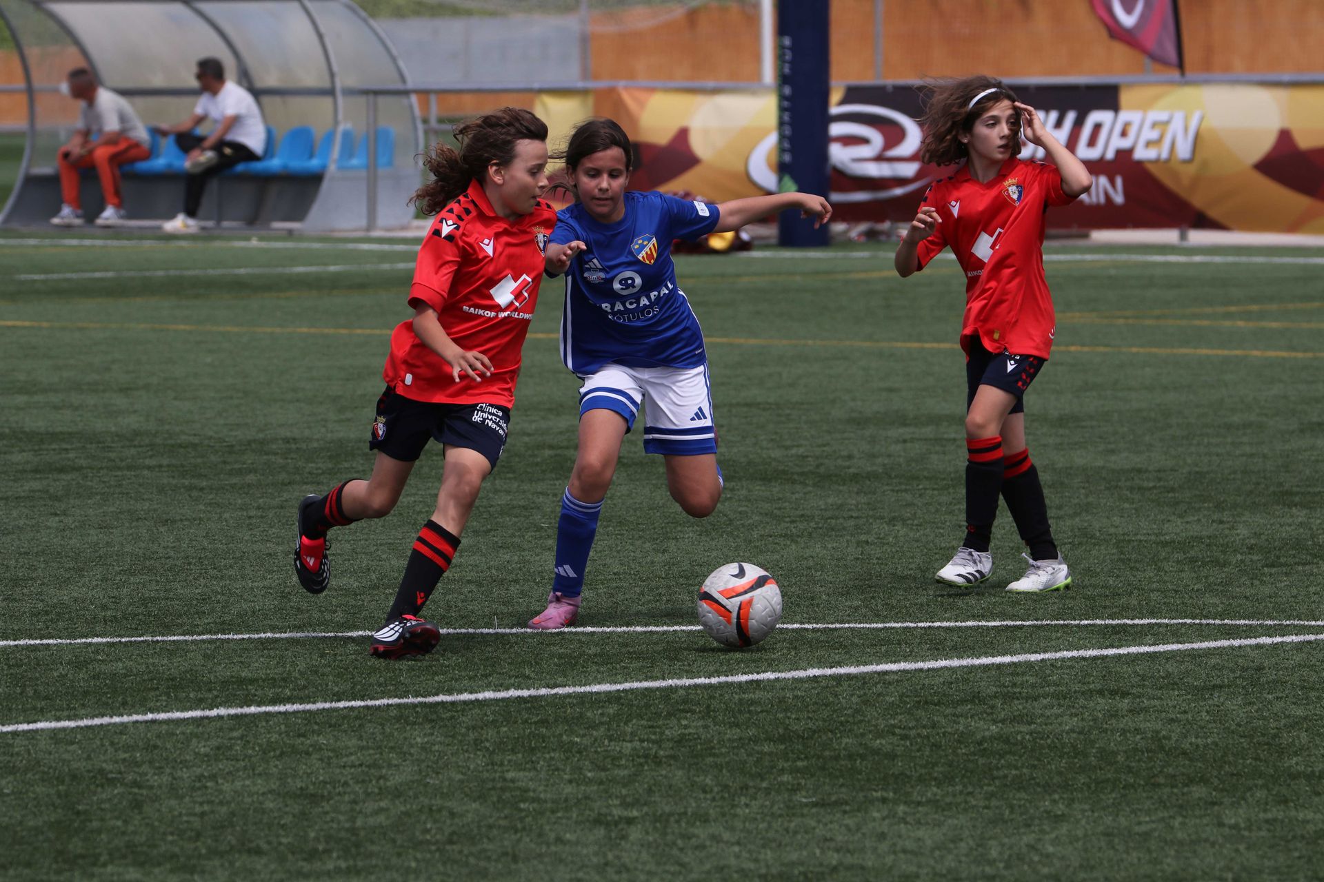 Three soccer players contest the ball on a turf field, two in red and one in blue.