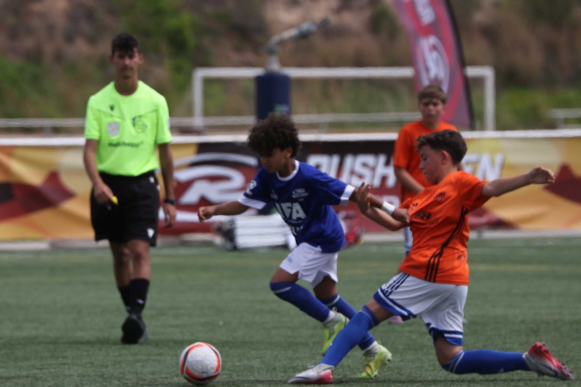 Youth soccer players in blue and orange contest the ball on a grass field with a referee in the background
