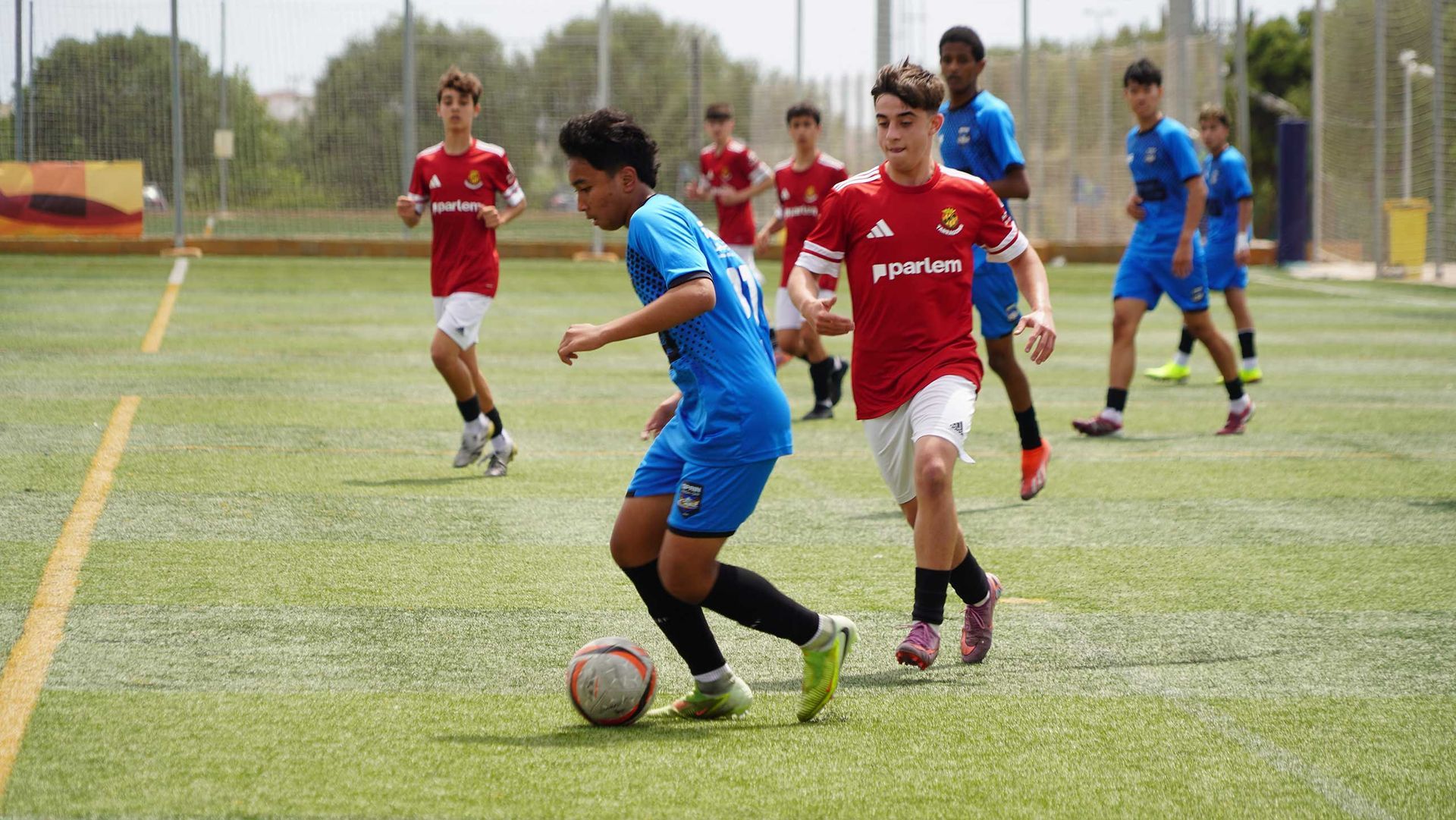 Soccer players in red and blue uniforms compete on a grassy field, one dribbling the ball in the foreground