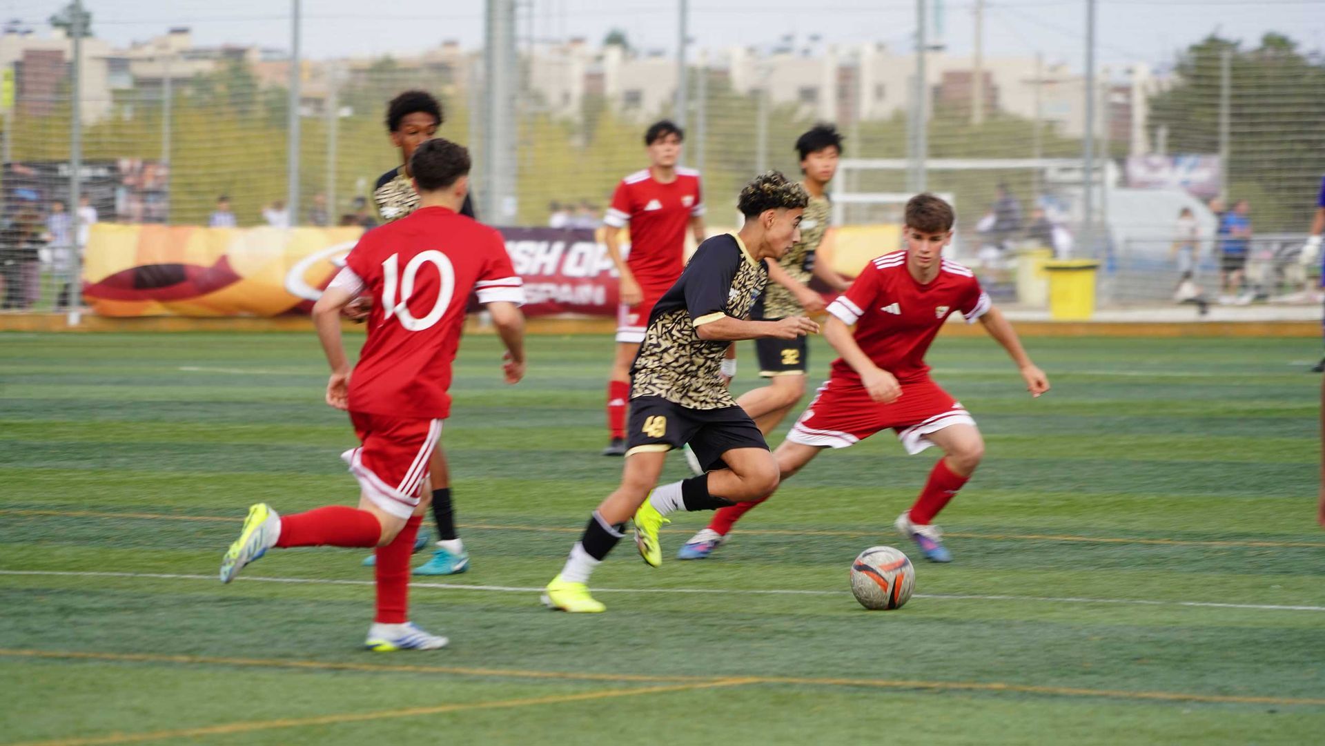 Soccer players in red and camo jerseys sprinting after the ball on a green field