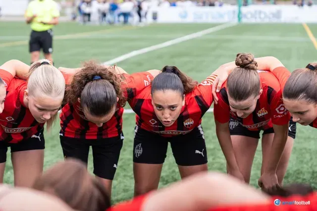 Female soccer team huddled together, wearing red and black jerseys, on a green field, focused expressions.