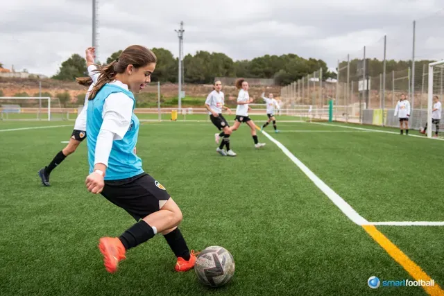 Woman in blue vest kicks soccer ball on green field, other players in background.