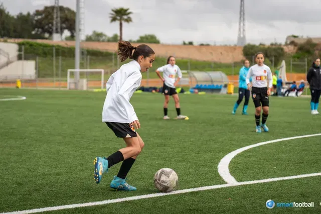 Girl in white soccer jersey dribbling a soccer ball on a green field, other players in the background.