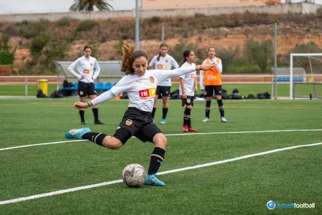 Woman in white and black soccer uniform kicking a ball on a green field. Other players in background.