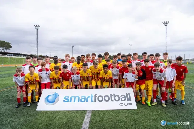 Youth soccer players in red/yellow jerseys pose with Smartfootball Cup sign on field.