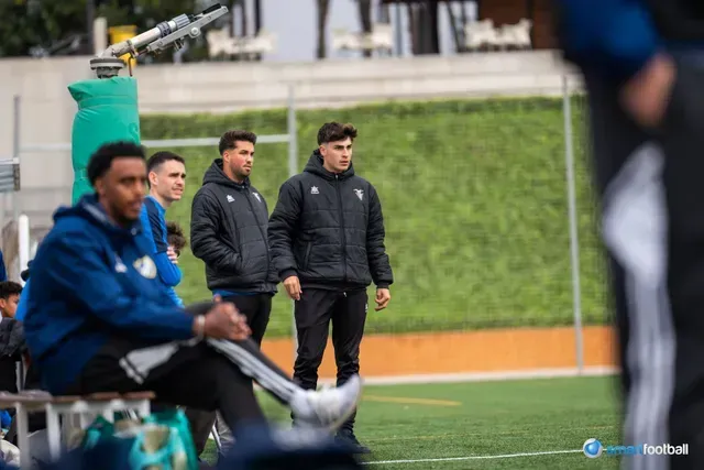 Soccer players on the sideline; man in black jacket and pants, others in blue, green netting in the background.