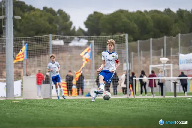 Boy in a white and blue soccer uniform dribbling the ball on a green field, others in the background.