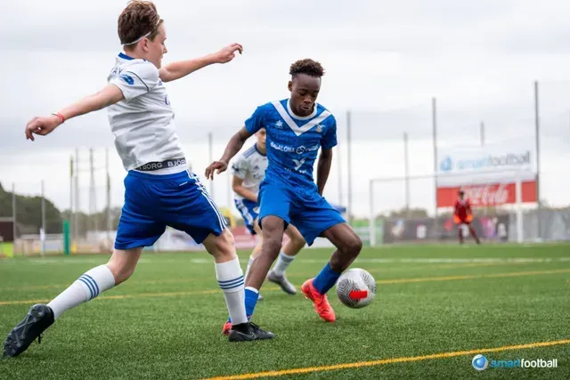 Two young soccer players in blue and white uniforms on a green field, one dribbling the ball.