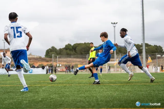 Soccer player in blue kicking ball on green field, two opponents run near, overcast sky.