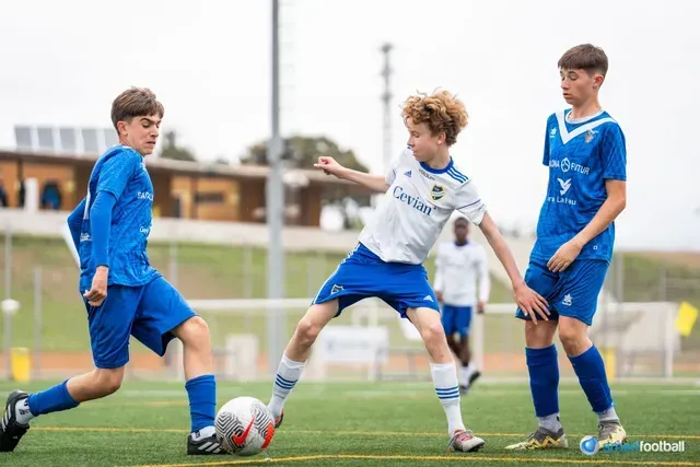 Three boys playing soccer on a green field, two in blue, one in white, kicking the ball.
