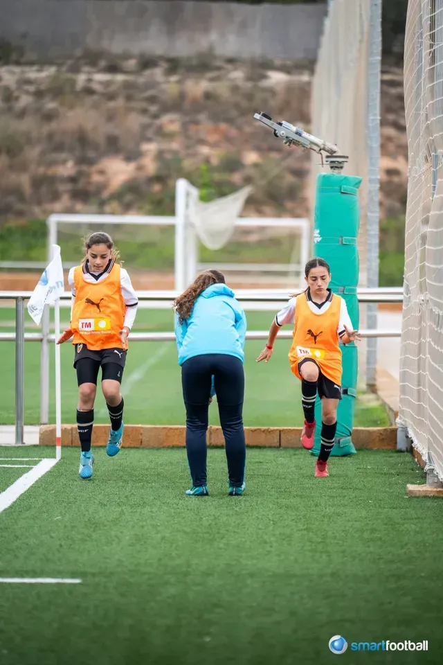 Two young soccer players in orange vests run drills with a coach on a field.