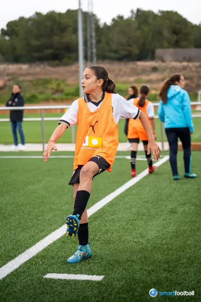 Girl in orange vest kicks a soccer ball on a green field, other players and spectators in the background.