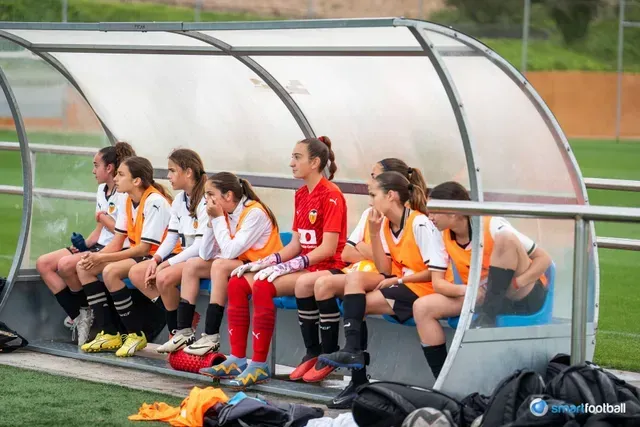 Soccer team on the bench, wearing orange/black uniforms, watching the game under a shelter.