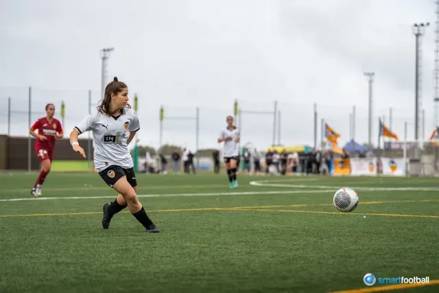 Female soccer player in white jersey dribbling ball on green field; others in the background.
