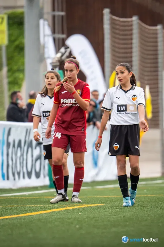 Soccer players on field; one in maroon uniform, two in white and black.