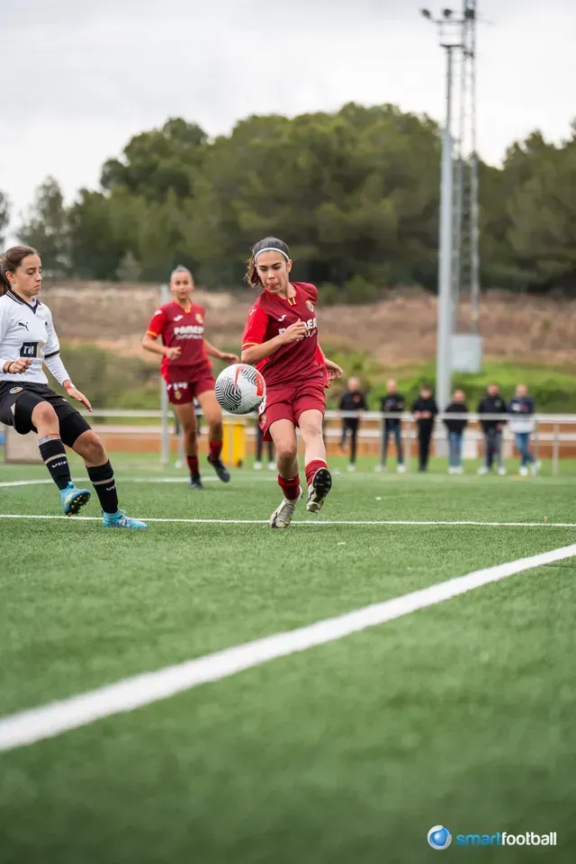 Soccer player in red kicking the ball on a green field, another player in black defending.
