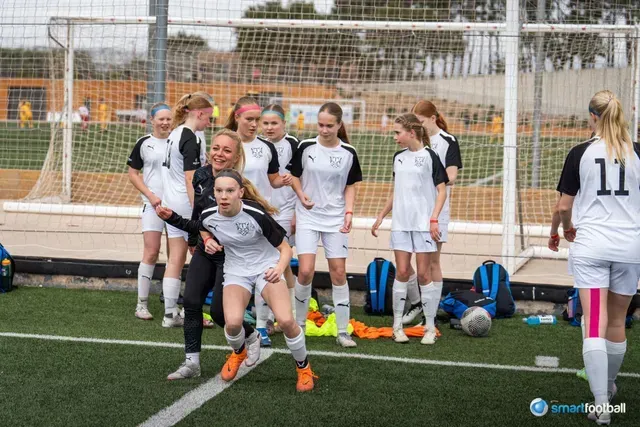 Girls' soccer team celebrating on a green field. Players in white and black uniforms cheer near a goal.