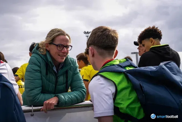 Woman in glasses talking to a young person. Others watch. Outdoors, overcast sky.