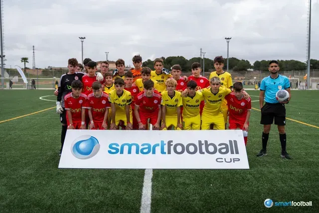 Soccer teams in red and yellow jerseys, posing with the Smartfootball Cup sign on a green field, referee present.