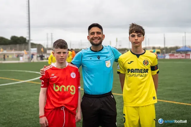 Three soccer players posing. Referee in turquoise smiles between players in red and yellow jerseys on a field.