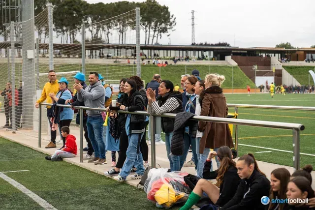 Fans watch a soccer game from behind a railing. People are cheering, and the setting is a sports field.