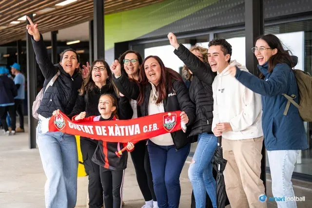 Group of fans cheer, holding a red and white banner for UD de Kativa C.F. Outdoors; excited expressions.